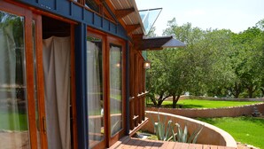 Une terrasse en bois avec des portes vitrées donnant sur une pièce, le tout entouré d’un jardin avec des arbres et une pelouse.