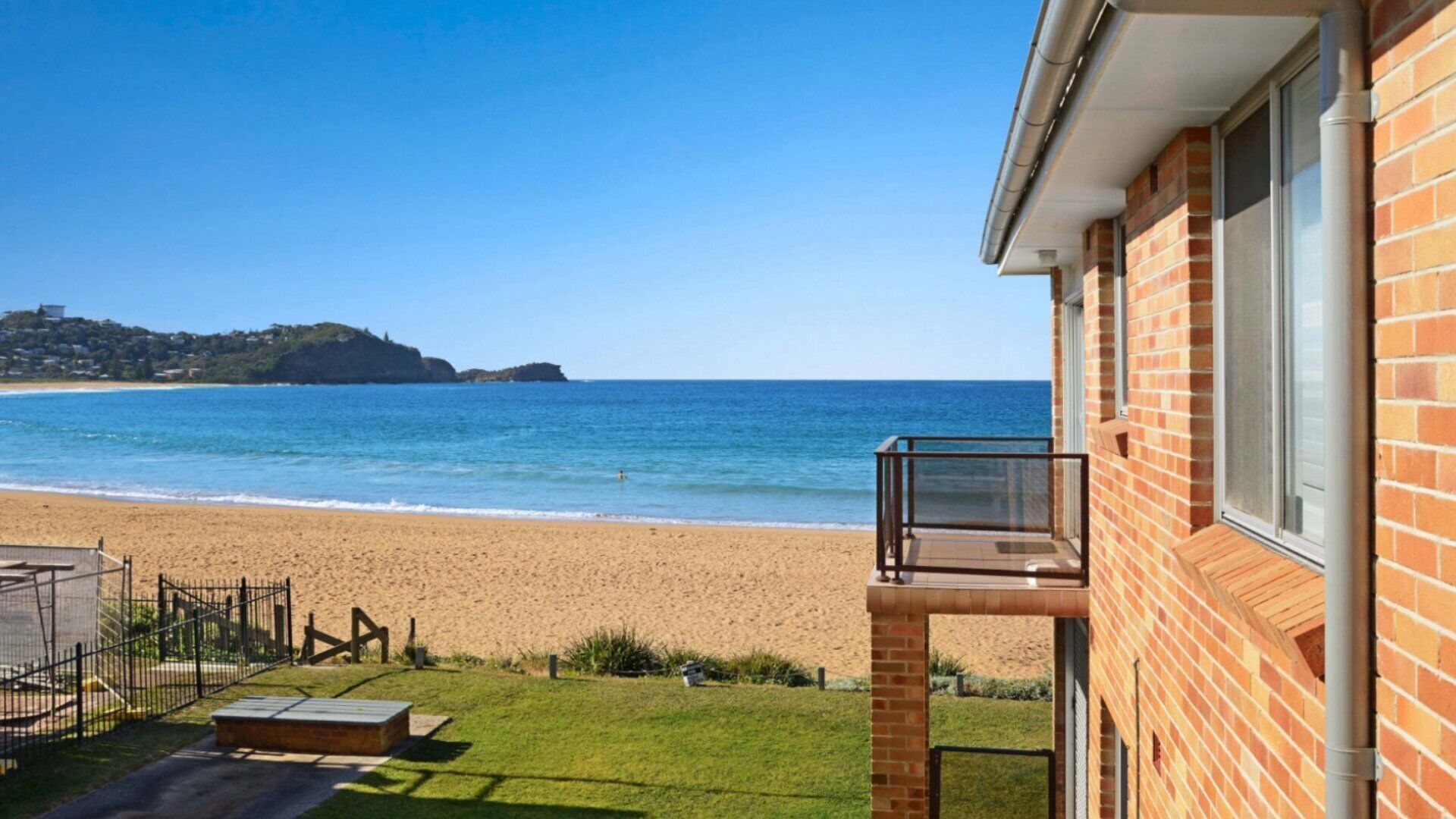 A beachfront property with a balcony overlooking the sea, a sandy beach, and a grassy area with a fence.