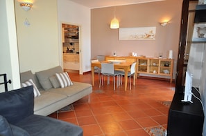 A living room with a sofa, dining area with chairs and a table, and a kitchen with cabinets and a hanging light fixture.