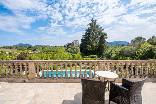 An outdoor patio with a stone balustrade, two wicker chairs, and a round table. In the background, there is a swimming pool and a view of a mountainous landscape.