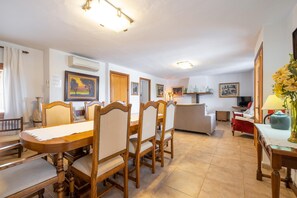 A dining area with a wooden table and chairs, a kitchen in the background, and a living area with a sofa and a TV.