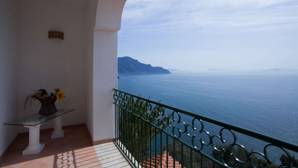 Ein Balkon mit Glastisch und Blumenvase, der Blick auf die Küste mit Meer und fernen Bergen freigibt.