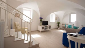 Ein geräumiger Wohnbereich mit Treppe, einem Fernseher auf einem Schrank, einem blauen Sofa und einer Lampe auf einem kleinen Tisch.