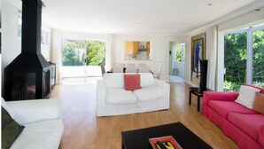 Een ruime woonkamer met een open haard, een witte bank, een rode bank en een keuken op de achtergrond.