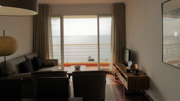 Hotelkamer met een groot raam met uitzicht op zee, een bank, een bijzettafeltje en een televisie op een houten standaard.