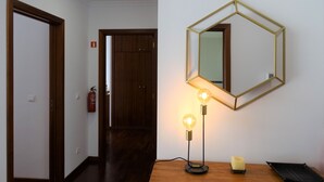 Een hal met een houten deur, een brandblusser, een houten vloer, een spiegel aan de muur, en een tafel met een lamp en een kaars.