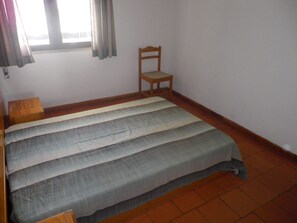 Um quarto pequeno e simples com uma cama, uma cadeira de madeira e uma janela com cortinas.