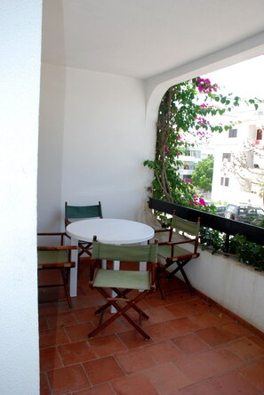 Uma pequena varanda com uma mesa redonda e duas cadeiras, uma parede branca e vista para o horizonte urbano através de uma janela com grade preta.