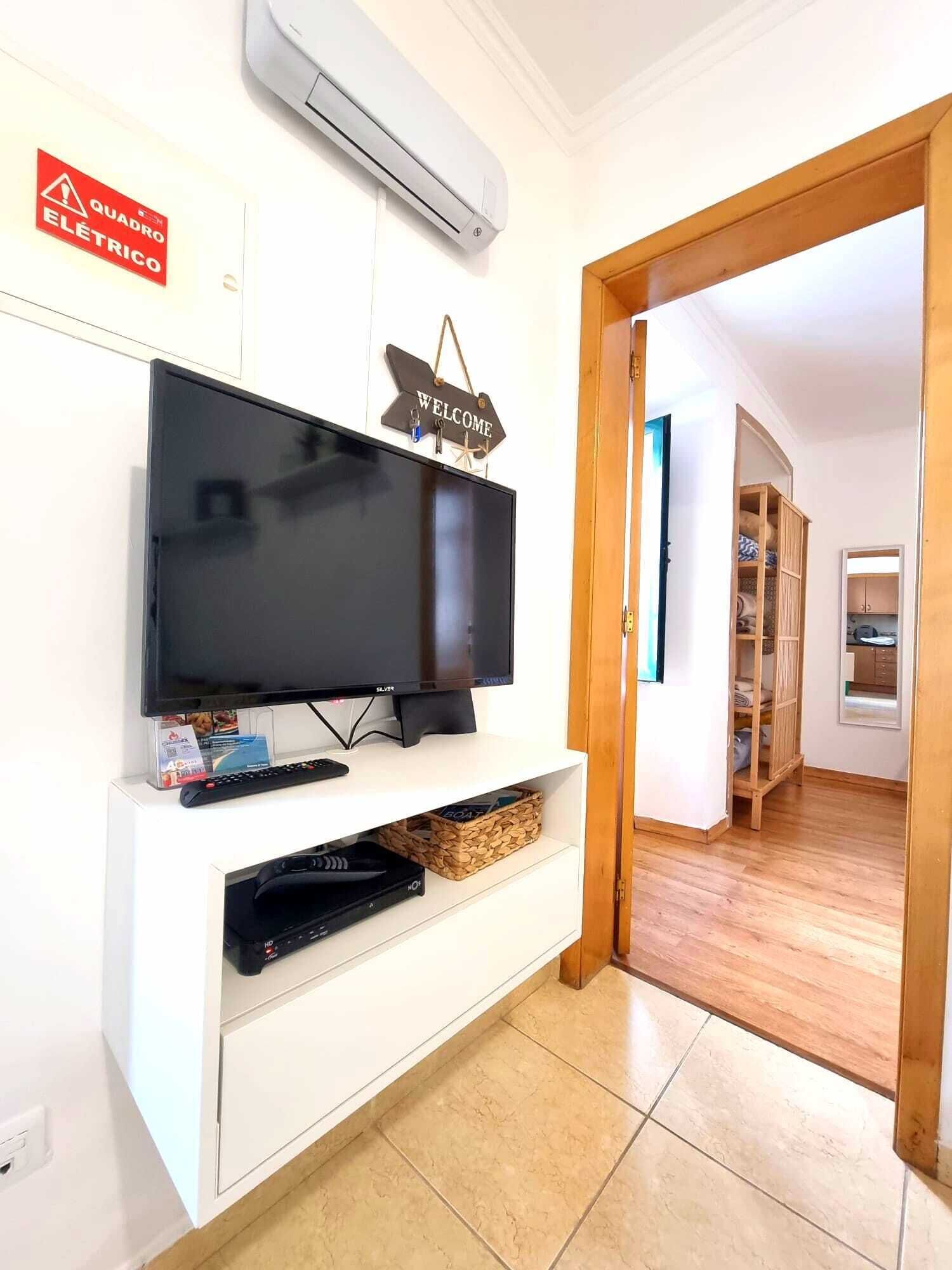 Una habitación con una televisión de pantalla plana sobre un pedestal blanco, un letrero de bienvenida arriba y un equipo de aire acondicionado en la pared.