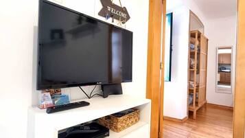 Una habitación con una televisión de pantalla plana sobre un pedestal blanco, un letrero de bienvenida arriba y un equipo de aire acondicionado en la pared.
