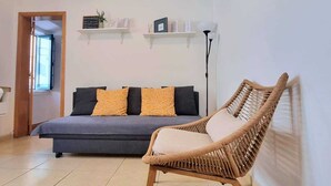 Een kamer met een bank, een rieten stoel en twee planken met spullen erop.