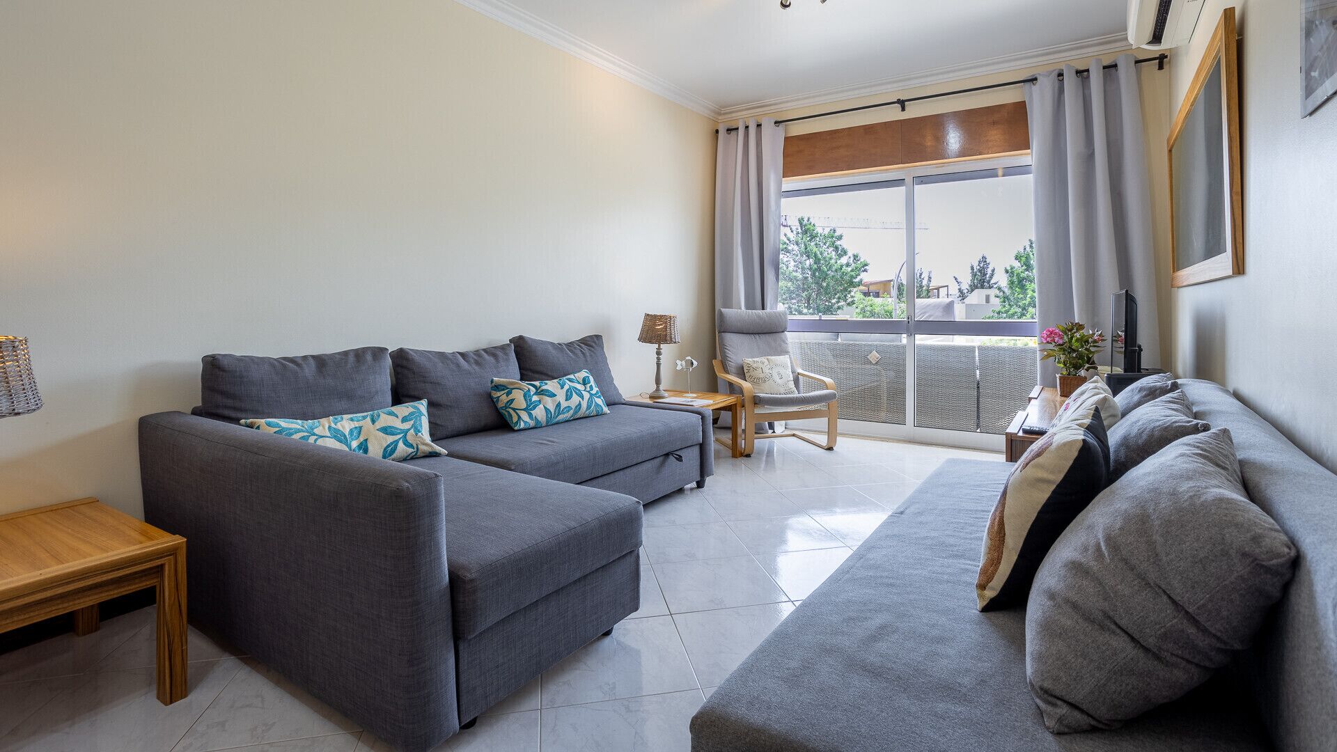 A modern living room with a grey sofa set, a wooden side table, and a balcony with curtains.