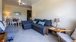 A modern living room with a grey sectional sofa, a wooden coffee table, and a side table with a lamp. There is a dining area with a white table and chairs in the background.