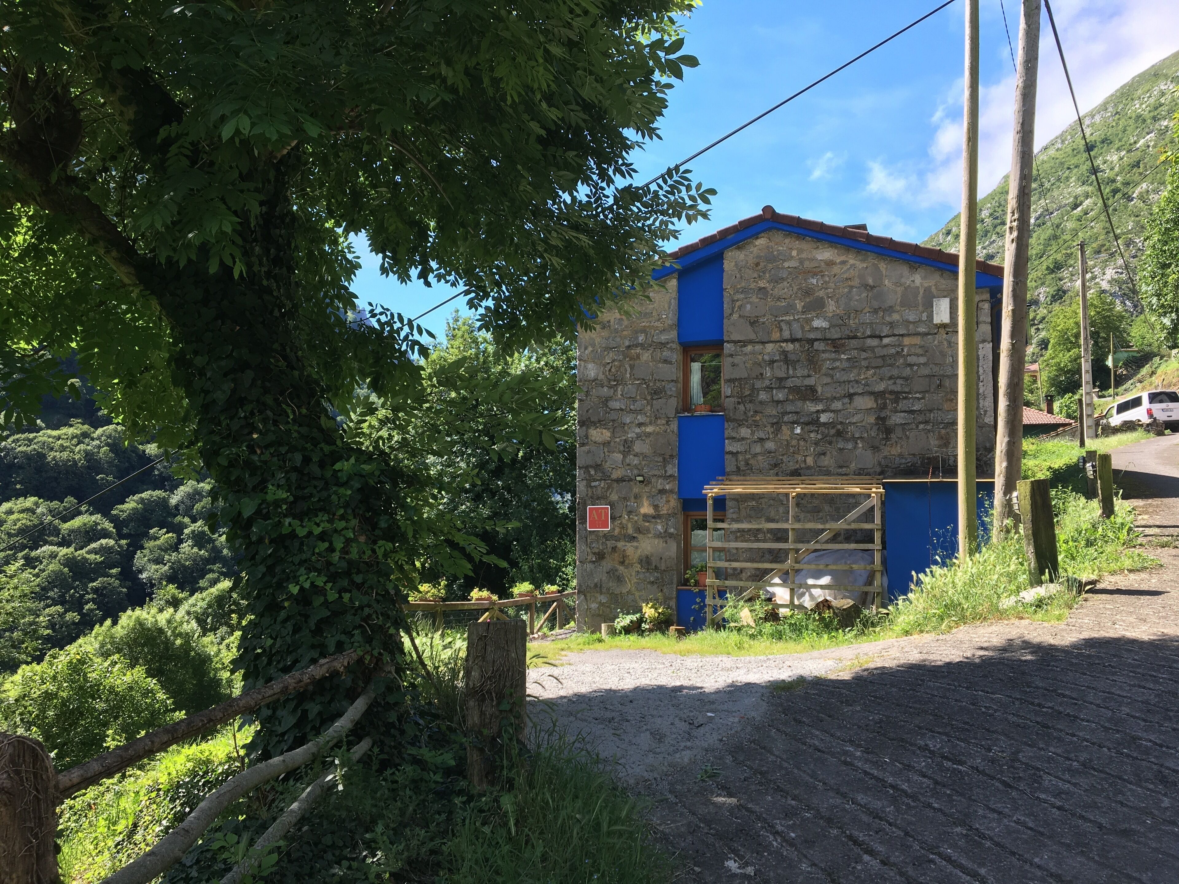 A rustic stone building with a blue door and a wooden gate, surrounded by lush greenery and a dirt path.