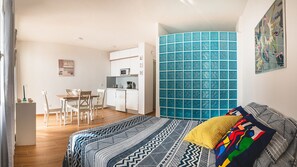 A compact hotel room with a bed, a small kitchenette, a dining area, and a bathroom with a blue tiled wall.