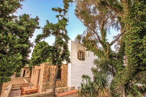 Un edificio de piedra con fachada blanca, una ventana pequeña y una puerta. Hay una mesa y sillas afuera, y un árbol en primer plano.