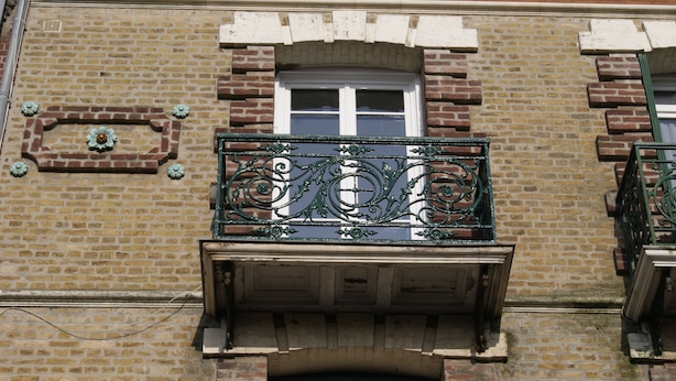 A building facade with a brick wall, a window with white trim, and a decorative wrought iron balcony railing. - Asylum Single Apartment (Mers-Les-Bains)