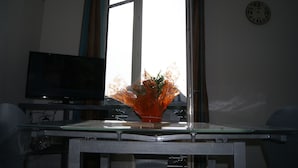 A room with a TV on a stand, a glass table with a vase, and a window with curtains. - Asylum Single Apartment (Mers-Les-Bains)