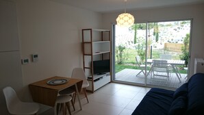 Un espace de vie compact comprenant un canapé, un téléviseur posé sur un meuble, une table à manger avec des chaises et une porte-fenêtre coulissante donnant sur une terrasse extérieure.