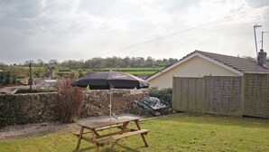 Una mesa de picnic de madera con un paraguas negro, un muro de piedra y una casa al fondo.