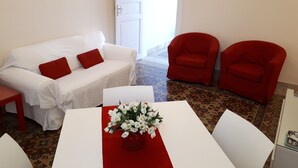 Una habitación con un sofá blanco, dos sillas rojas, una mesa blanca con camino de mesa rojo y un jarrón con flores blancas.