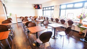 Une salle de classe avec des pupitres et des chaises en bois, un téléviseur fixé au mur et de grandes fenêtres.