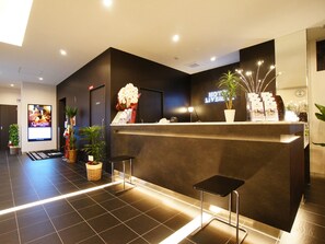 A hotel reception area with a modern design, featuring a reception desk, potted plants, and a digital screen displaying hotel information. - HOTEL LiVEMAX Himeji Shiyakusho Mae (Himeji)