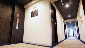 Un couloir d’hôtel avec un sol recouvert de moquette, une pancarte indiquant la chambre 6F et une porte dotée d’un bouton d’appel d’ascenseur.