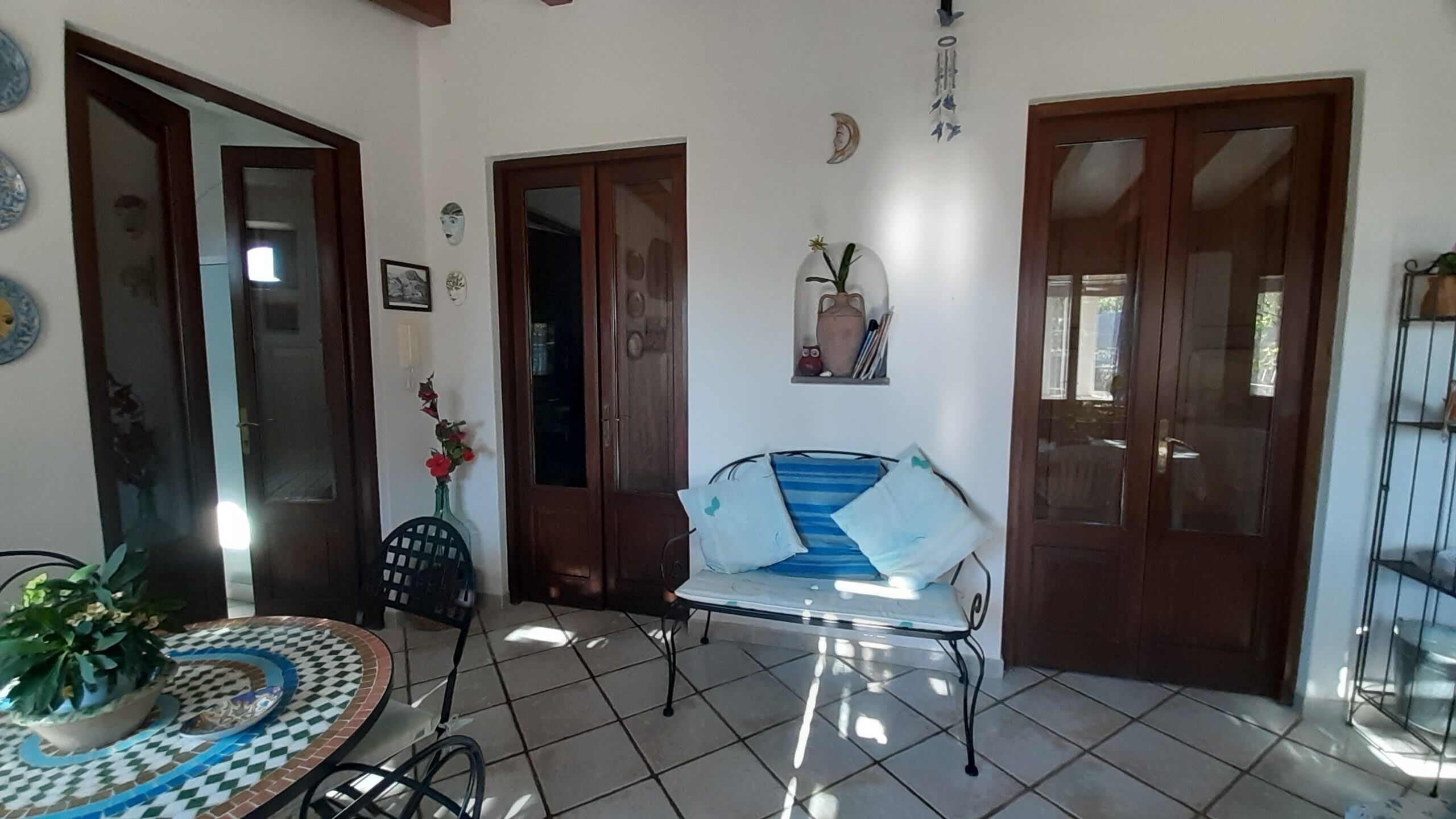 Una stanza con pavimento piastrellato, un tavolo rotondo con tovaglia fantasia, una sedia a righe blu e bianche e una pianta in vaso.