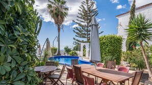 An outdoor patio with a wooden table and chairs, a swimming pool, and various plants and trees.