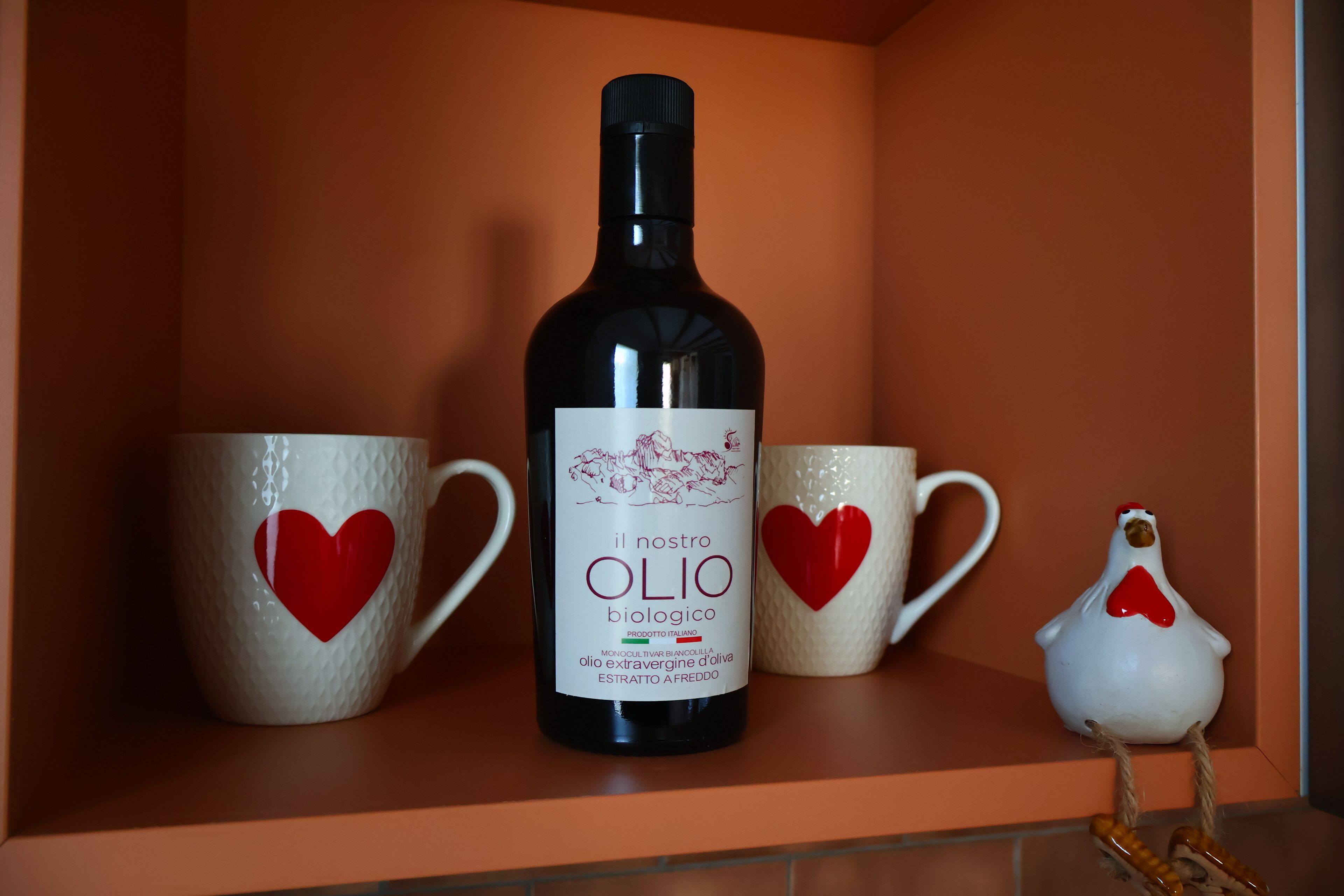 Eine Flasche 'il nostro OLIO biologico' Wein, zwei Keramiktassen mit Herzmotiven und eine Keramik-Hühnchenfigur auf einem Regal.