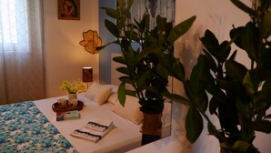 A neatly made bed with a floral bedspread, a bedside table with a vase of flowers, a framed picture on the wall, and a large plant in the foreground.