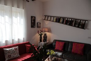 Una sala de estar con un sofá rojo, un sofá de cuero negro, una estantería, una lámpara y una ventana con cortinas de gasa.