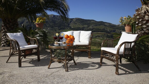 A patio with wicker furniture, a table with a fruit basket and drinks, and a view of hills in the background.