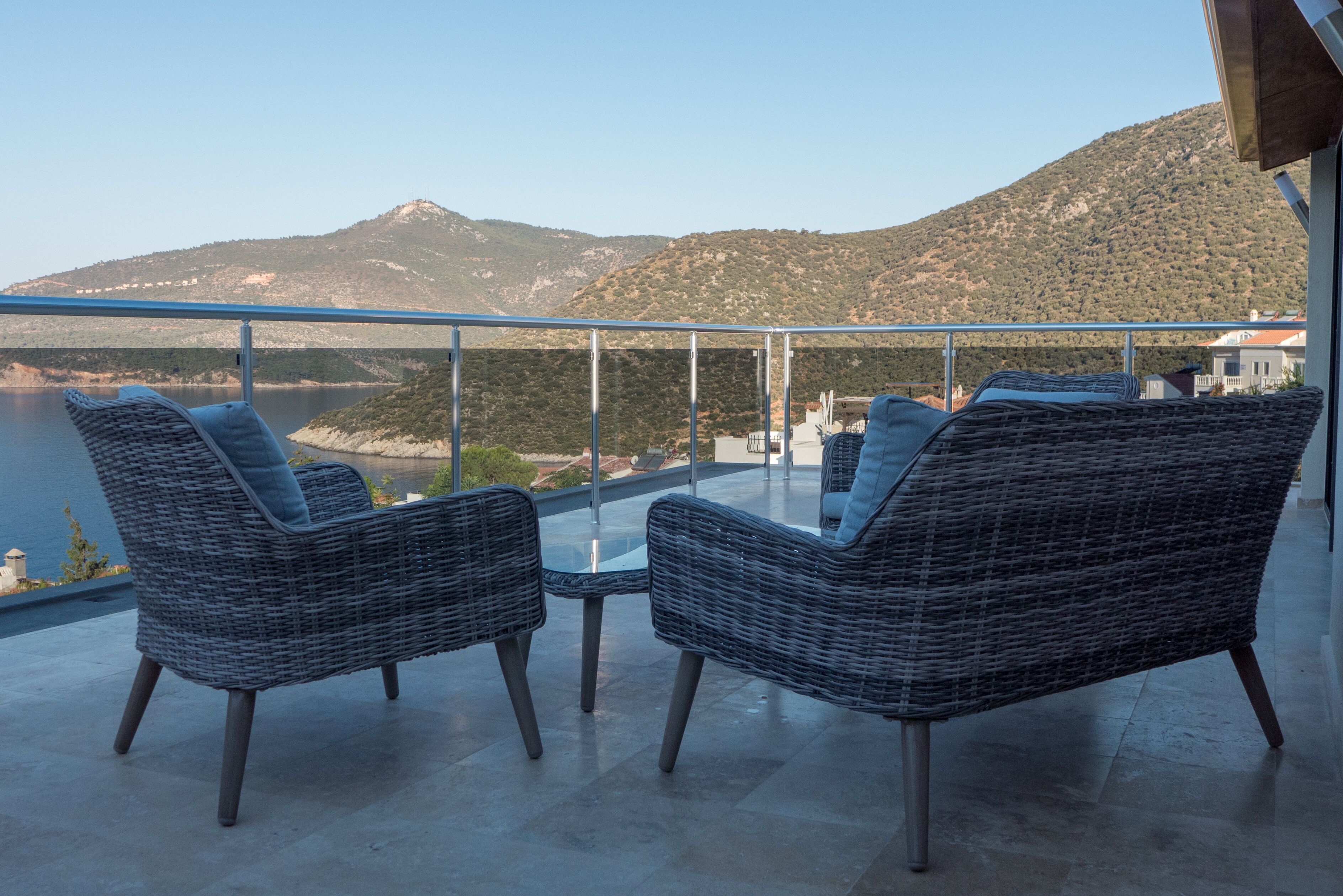 A balcony with wicker chairs and a glass table overlooking a mountainous landscape.