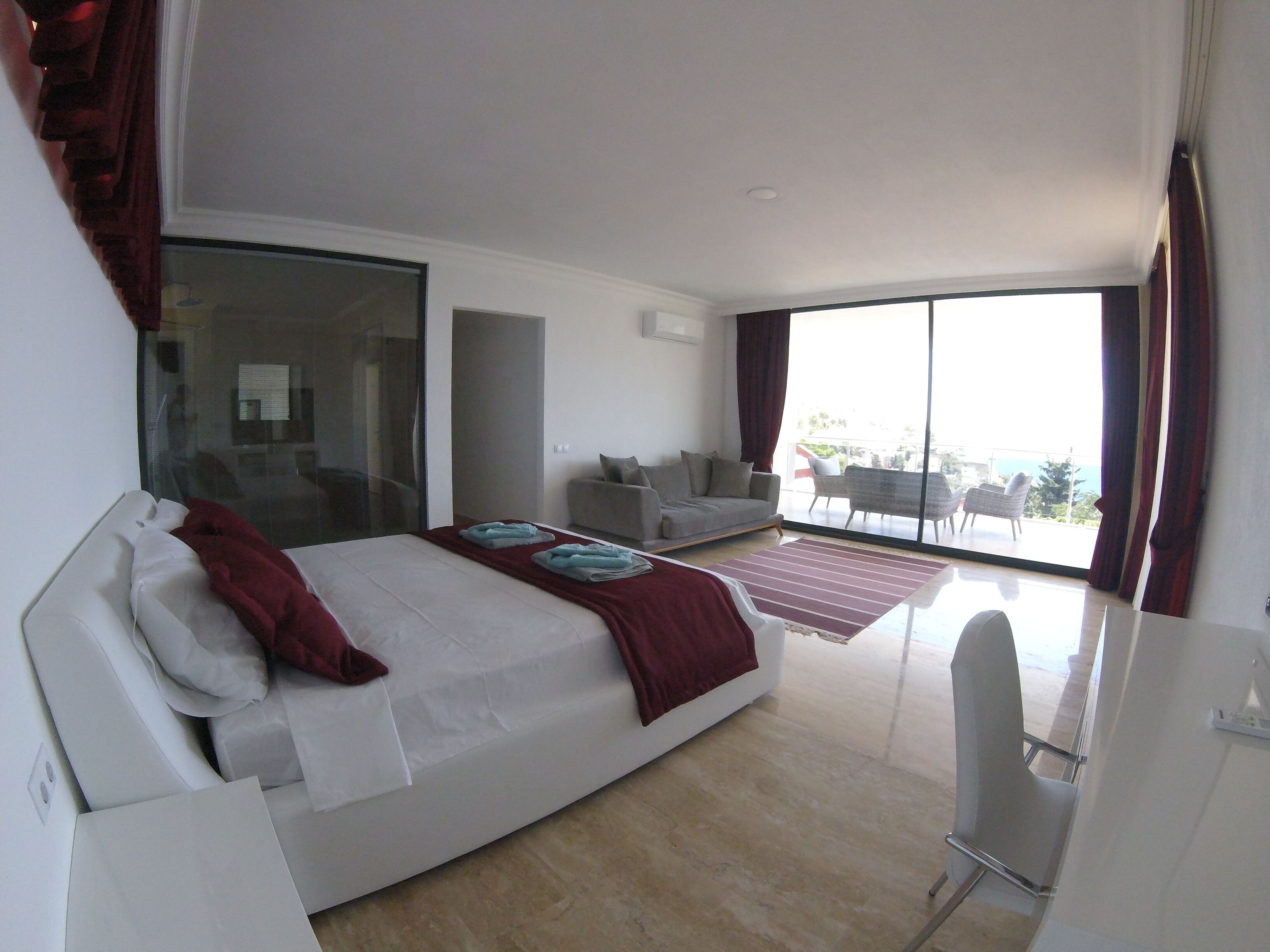 A modern hotel room with a large bed, a sitting area with a sofa and armchairs, a balcony with a view, and a desk with a chair.