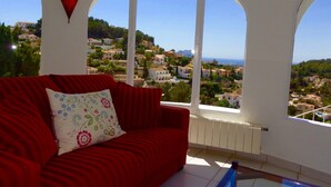 Un divano rosso con un cuscino floreale, un lampione rosso e vista su una località costiera attraverso ampie finestre ad arco.