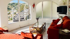 Un soggiorno con un divano rosso, un tavolino in vetro e una TV su un mobile bianco. Si vede un'ampia finestra con vista su un ponte e sulla vegetazione.