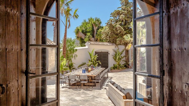 An open wooden door leading to a patio with a table set for dining, surrounded by greenery and a white wall.