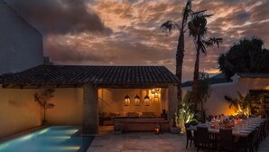 A poolside dining area with a long table set for guests, a pool, and a covered seating area with ambient lighting.