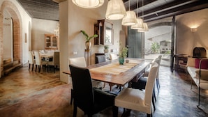 A spacious dining area with a long wooden table, multiple chairs, hanging light fixtures, and a view of the outside through large windows.