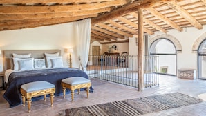 A bedroom with a wooden ceiling, a bed with a blue bedspread, two upholstered chairs, and a view of the outside through an arched window.