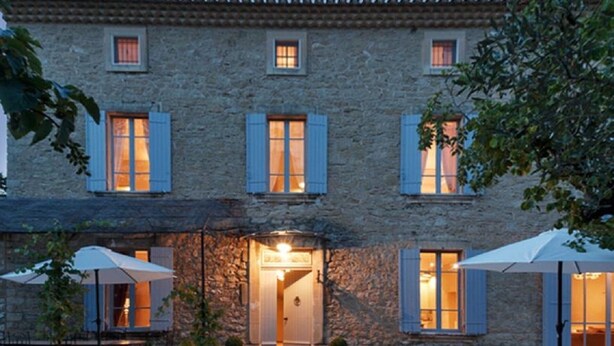 Ein Gebäude aus Stein mit vielen Fenstern, von denen einige mit Fensterläden versehen sind, und einer zentralen Tür. Im Vordergrund befinden sich Sonnenschirme und ein Baum.