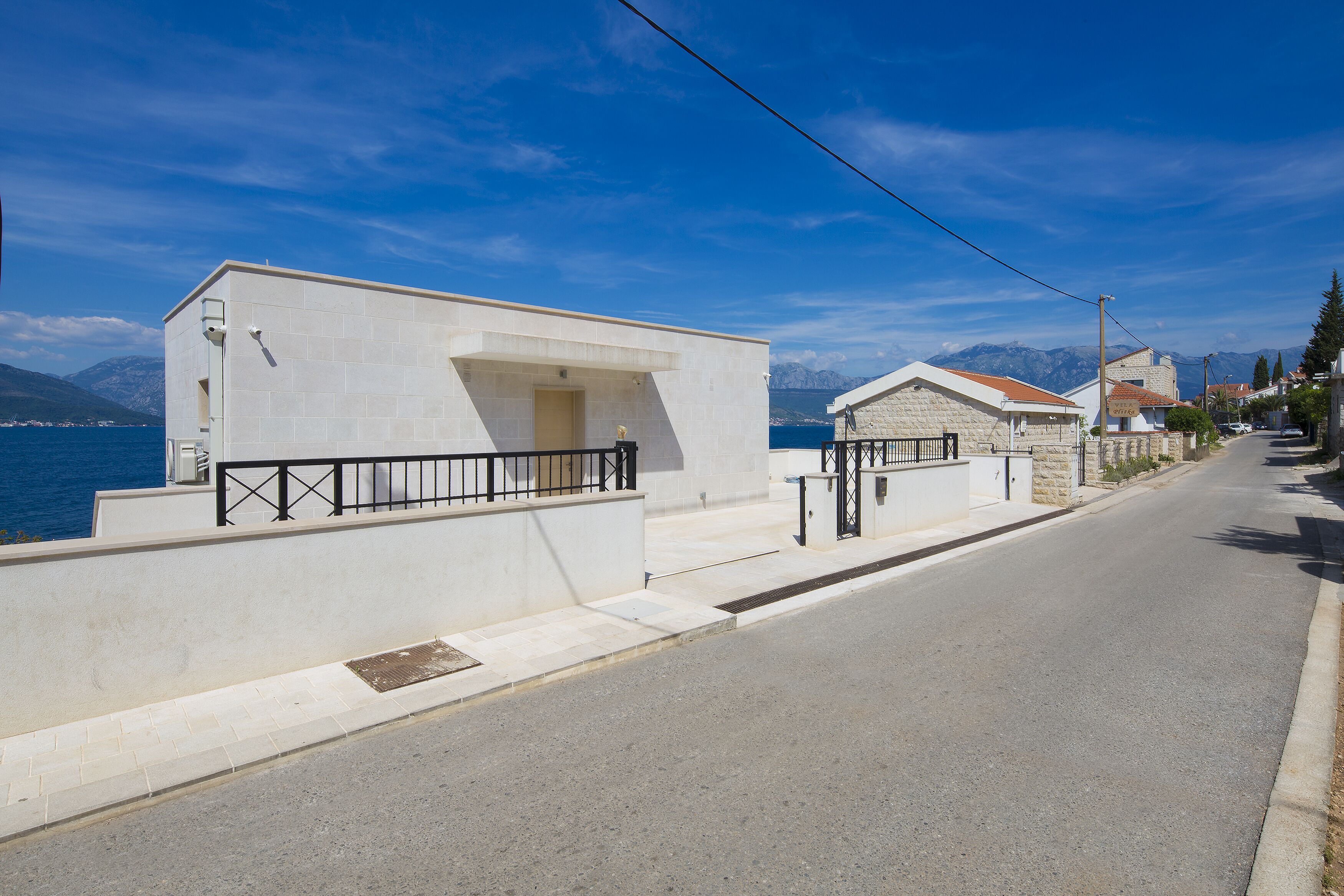 Une rue côtière bordée de bâtiments blancs, sous un ciel bleu azur, avec une vue sur la mer et les montagnes en arrière-plan.
