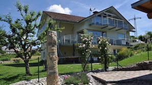 A two-story house with a balcony, surrounded by a garden with flowers and a stone pillar.