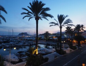 A marina at dusk with boats docked, palm trees, and a road with pedestrians.