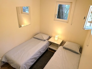 A small, single-room accommodation with two beds, a bedside table with a lamp, and a window with a view of trees.