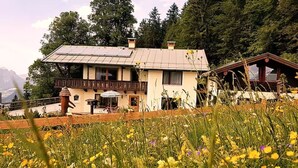 Une maison avec un balcon, un jardin parsemé de fleurs sauvages et une vue sur les montagnes en toile de fond.