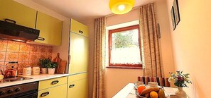 Une cuisine avec des meubles de couleur jaune, une cuisinière, une fenêtre avec des rideaux et un panier de fruits posé sur le comptoir.