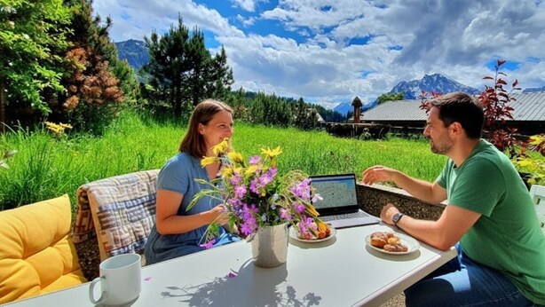 Deux personnes sont assises à une table à l’extérieur, devant un ordinateur portable, entourées de verdure et avec des montagnes en arrière-plan.